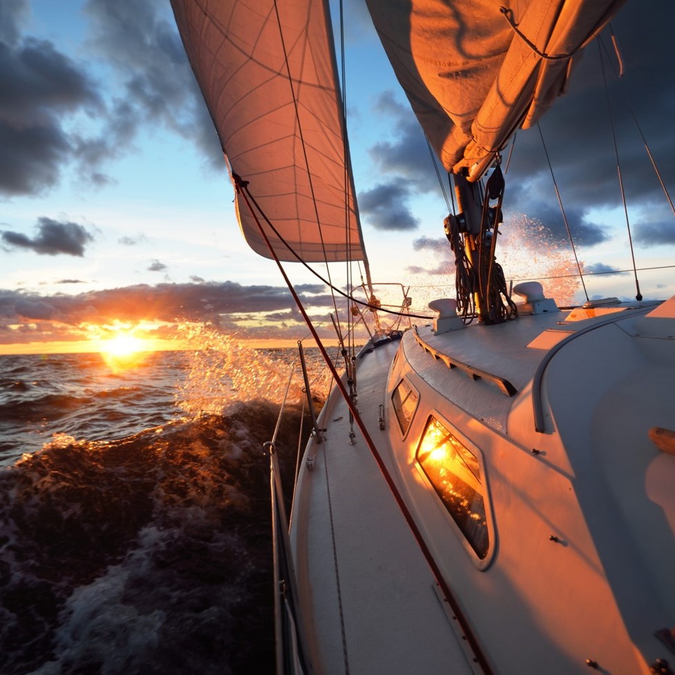 A sailboat navigating ocean waves at sunset, with water splashing.