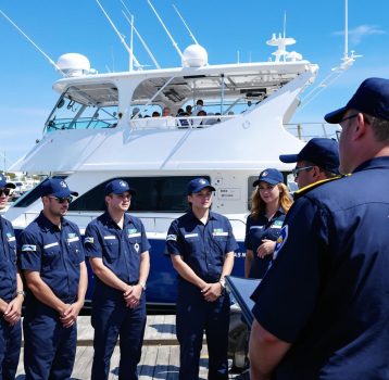 A boat crew in navy uniforms standing in formation while receiving instructions from a supervisor.