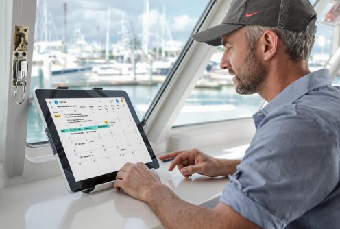 A man reviews a saltwater boat engine maintenance schedule on a tablet inside a boat cabin with a marina in the background.