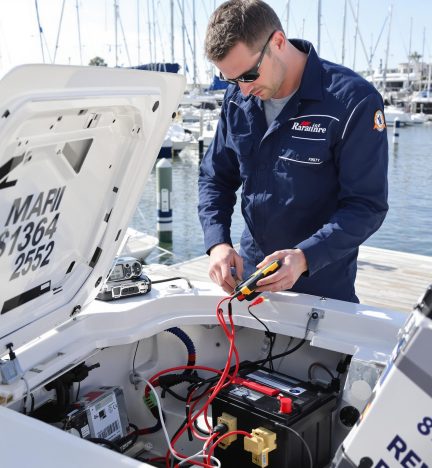 A marine technician in blue coveralls uses a multimeter to conduct boat battery testing and replacement at the marina.