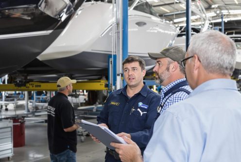 A boat technician in a navy uniform discusses repair details with two clients inside a professional boat repair facility. They are reviewing documents while multiple boats on lifts and various tools are visible in the background.