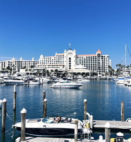 A vibrant marina scene at Loews Coronado Bay Resort, featuring yachts and boats docked in the calm waters. The grand resort building with Mediterranean-style architecture and palm trees is visible in the background under a bright blue sky.
